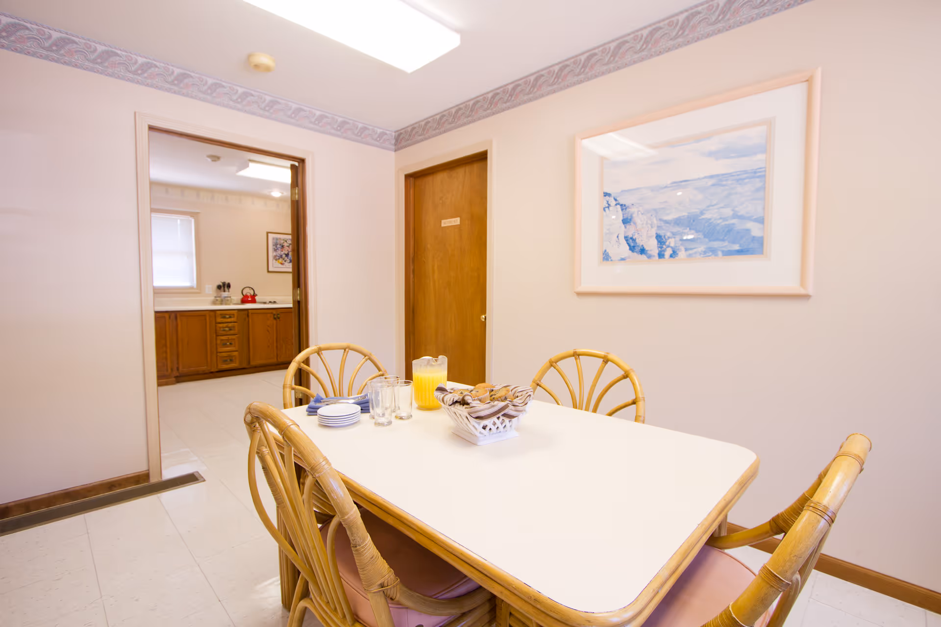 A small dining area with a rectangular white table surrounded by four wooden chairs with woven backs. On the table, there is a basket of bread rolls, a pitcher of orange juice, several glasses, and a stack of plates. The room has light-colored walls with a framed picture of a landscape hanging on one wall. An open doorway leads to a kitchen area with wooden cabinets and a red kettle on the stove.