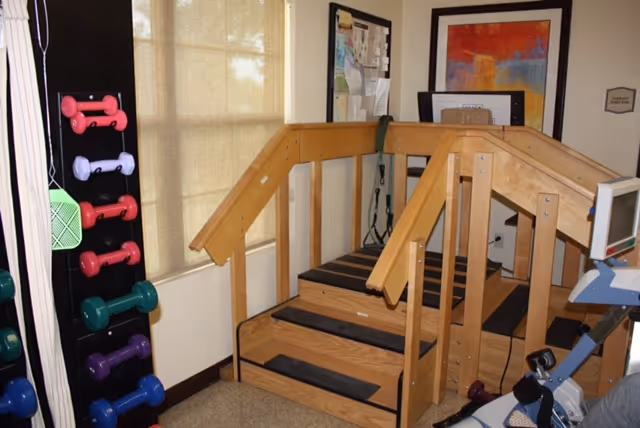 Indoor physical therapy area with wooden stairs equipped with handrails on both sides, a rack of colorful dumbbells on the left, and exercise equipment partially visible on the right. The room has beige walls and windows with blinds.