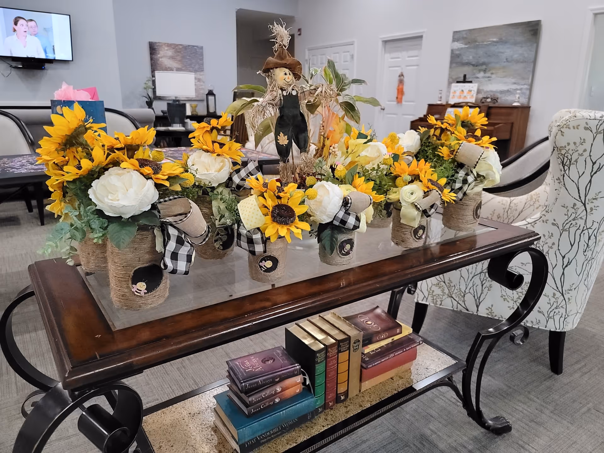 A cozy living room area with a glass-top wooden coffee table decorated with multiple small flower arrangements featuring sunflowers, white roses, and checkered ribbons. A small scarecrow figurine is placed among the flowers. Below the table, there are several stacked books. In the background, there are upholstered chairs with a tree branch pattern, a piano, a wall-mounted TV, and various decorative items.