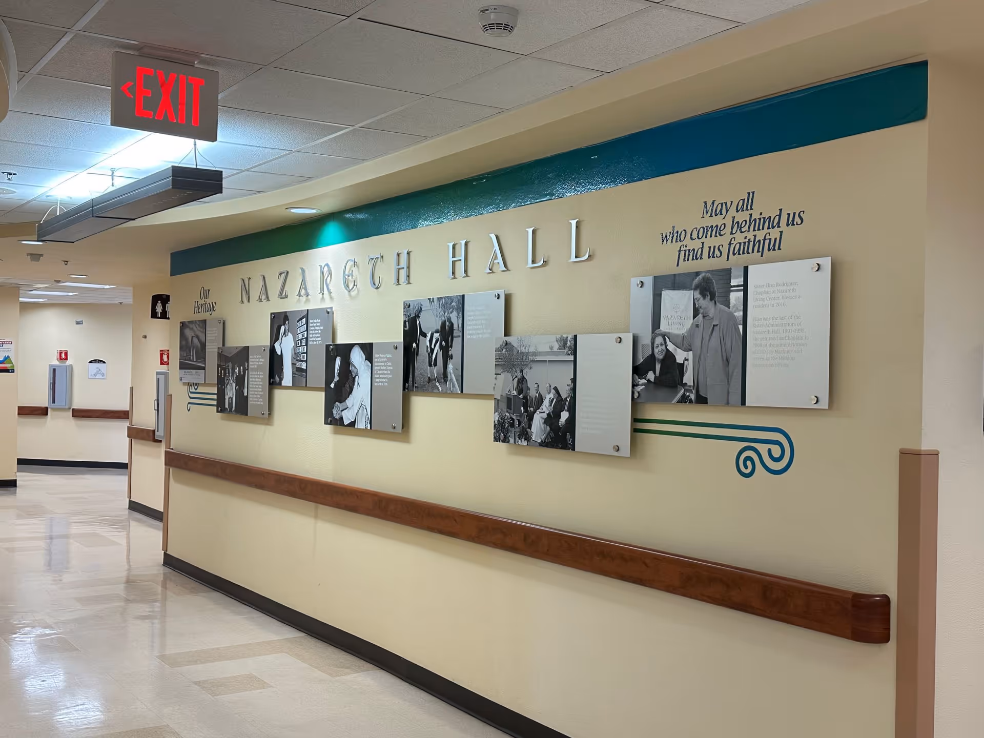 Interior hallway of Nazareth Hall with wall-mounted photos and signage, handrails, and an illuminated exit sign.