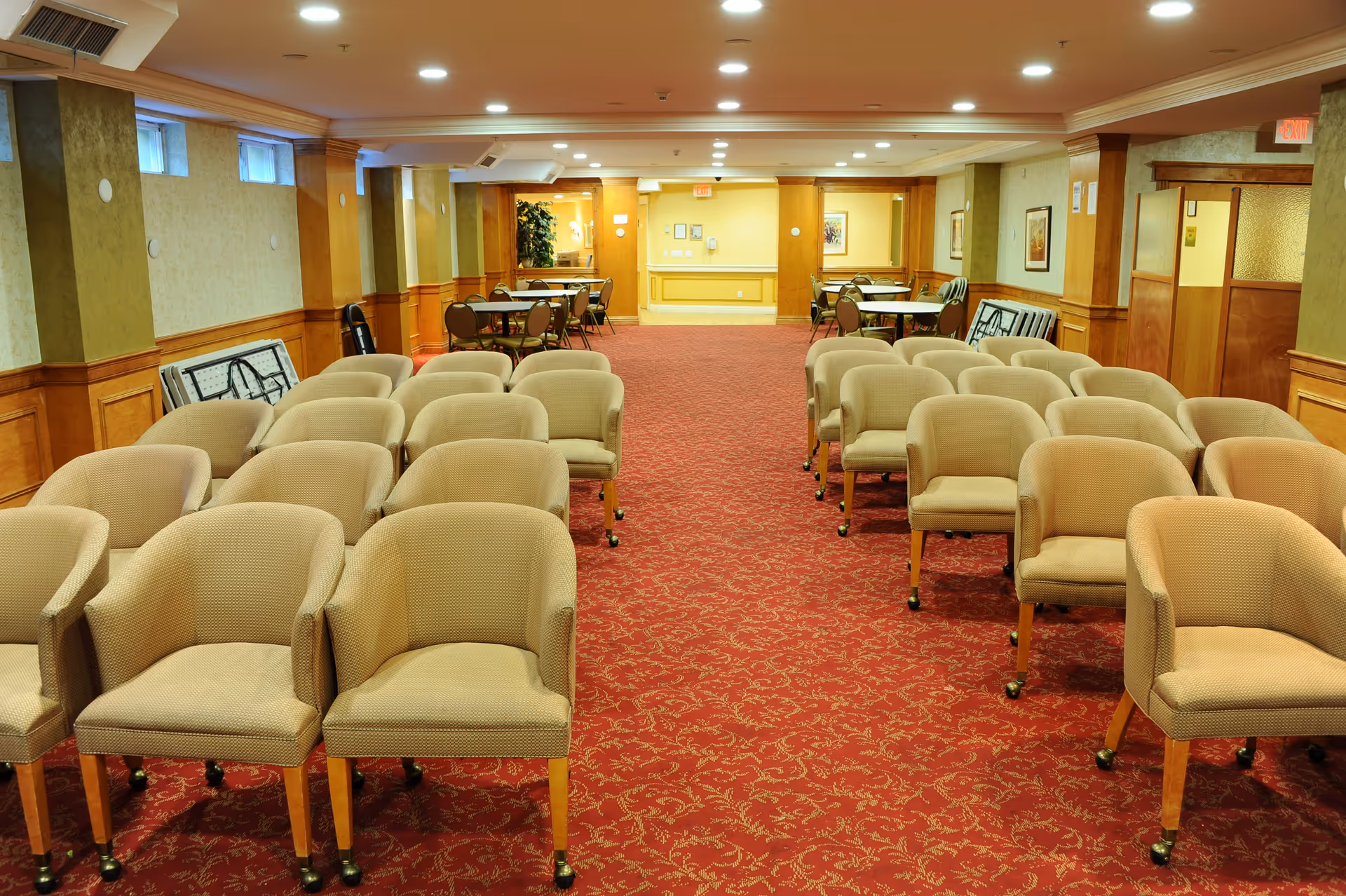 Carpeted communal room with rows of beige upholstered chairs arranged in two columns and tables near the back.