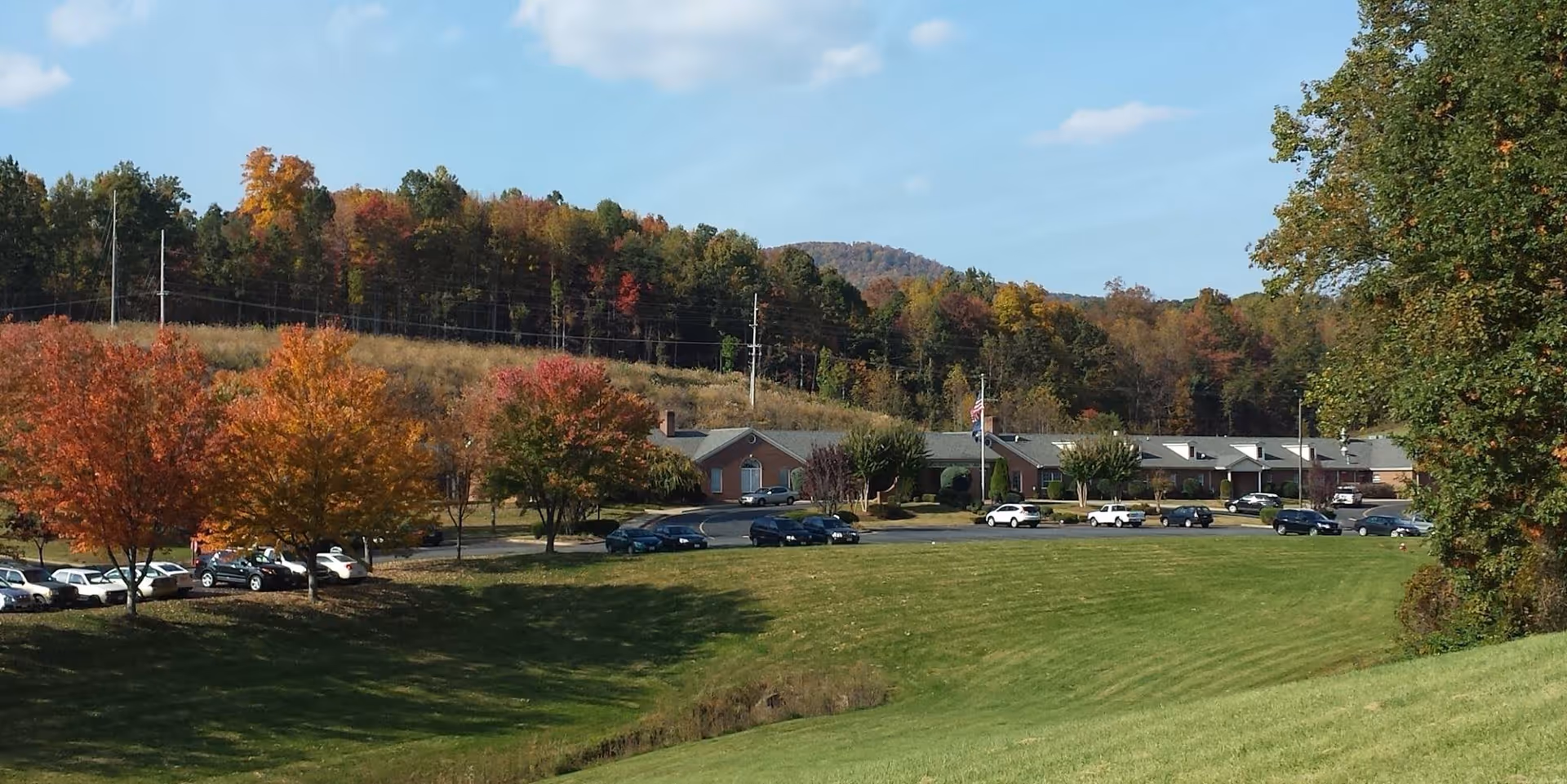 Wide exterior view of The Landmark Center facility with a long single-story building surrounded by a parking lot with several cars. The scene includes green grass, trees with autumn foliage, and forested hills in the background under a clear blue sky.