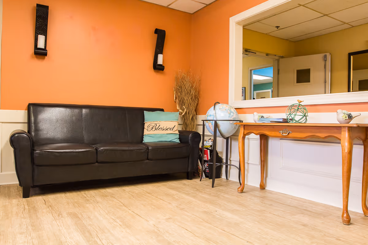 A cozy interior room with an orange accent wall featuring two black wall-mounted candle holders. There is a black leather couch with a decorative pillow that says 'Blessed'. Next to the couch is a globe on a stand and a wooden table with decorative items including a green spherical sculpture and small bird figurines. A large window or mirror is set in the wall above the table, reflecting part of the adjacent room.