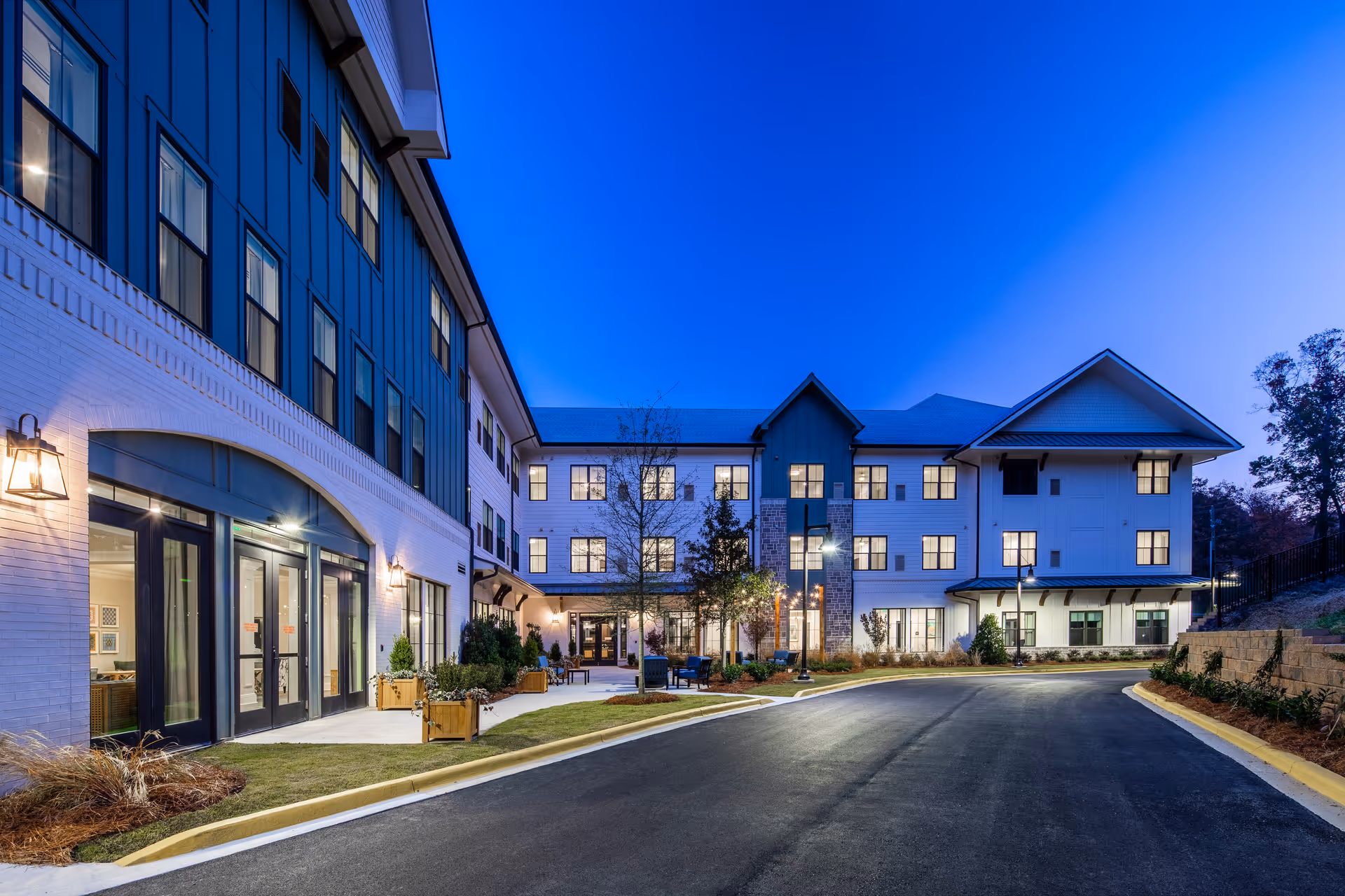 Front exterior of a multi-story senior living building at dusk with illuminated windows, entrance, and a curved driveway.