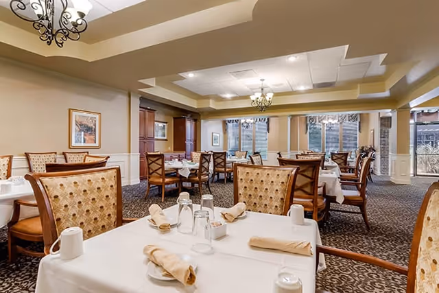A dining room in a senior living facility with multiple tables covered in white tablecloths, each set with beige napkins, glasses, and cups. The room features patterned carpet, upholstered chairs, chandeliers, and large windows letting in natural light.