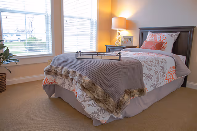 A neatly made single bed with patterned white and orange bedding and a gray throw blanket with a fur trim in a well-lit bedroom. There is a bedside table with a lamp and a framed picture, and two large windows with white blinds letting in natural light. A potted plant is visible near the window.