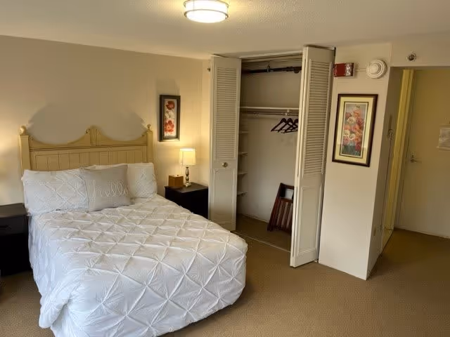 A neatly made bed with a white quilt in a furnished bedroom featuring an open closet, nightstands, a lamp, and framed artwork.
