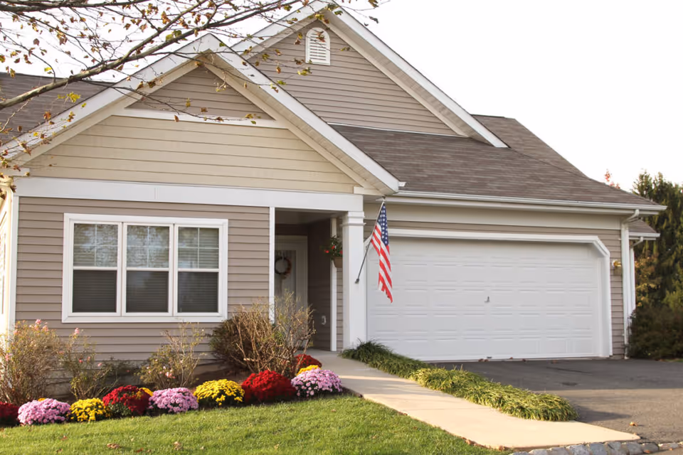 Front exterior of a single-story house with an attached two-car garage, American flag, and flower beds.