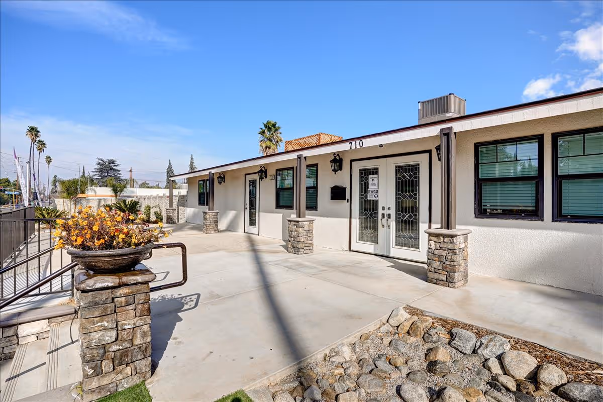 Front exterior of a single-story senior living building showing a concrete entry patio, double glass doors, stone columns and planters.