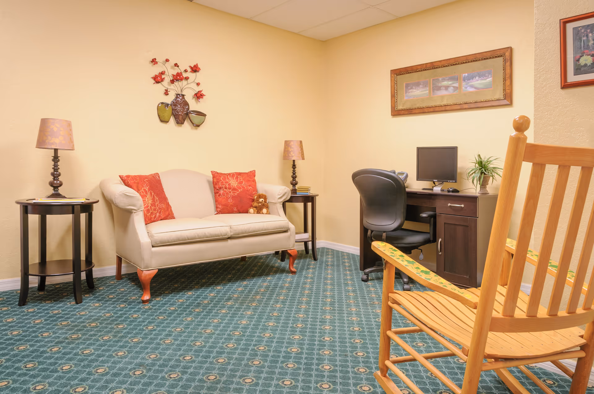 A cozy room with a beige loveseat adorned with two red floral pillows and a small teddy bear, flanked by two dark wooden side tables each with a lamp. There is a wooden rocking chair in the foreground and a dark wooden desk with a computer and a plant against the wall. The walls are painted light yellow and decorated with framed pictures and a floral wall ornament. The floor is covered with a green patterned carpet.