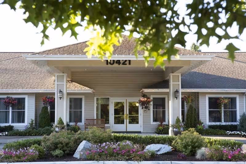 Front exterior view of a single-story building with a peaked roof, white columns, and double glass doors. The building is surrounded by well-maintained landscaping including bushes, flowers, and hanging flower baskets. The address number 10421 is visible above the entrance. Green tree leaves partially frame the top of the image.