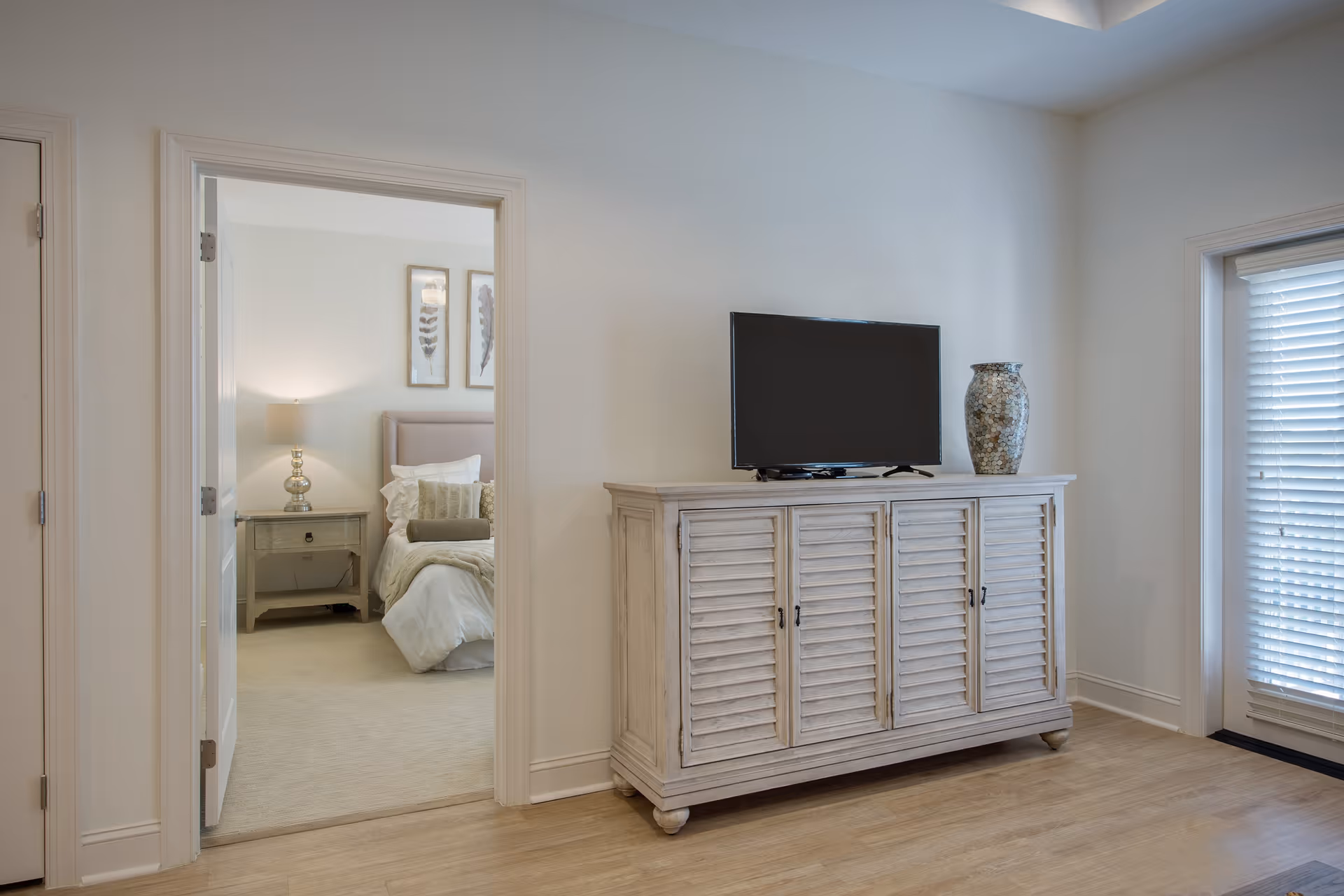 Light, neutral living area with a TV on a shutter-front cabinet, a doorway showing a bedroom, and window blinds.