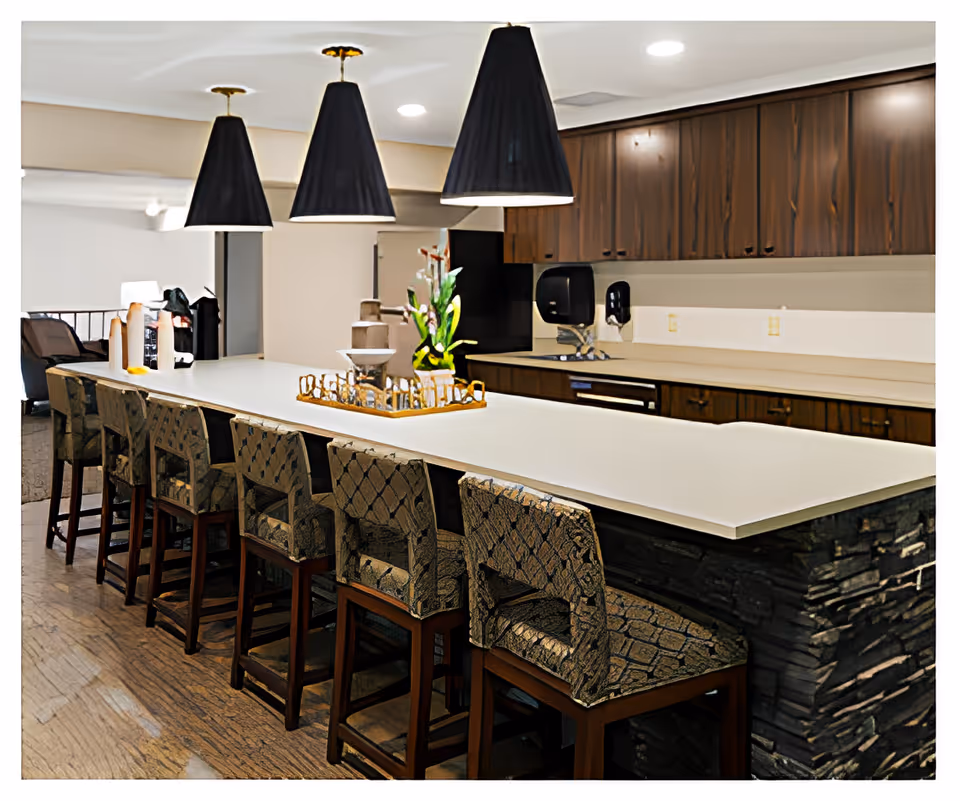 Long communal kitchen island with bar stools, pendant lights, and wood cabinets in a senior living facility.