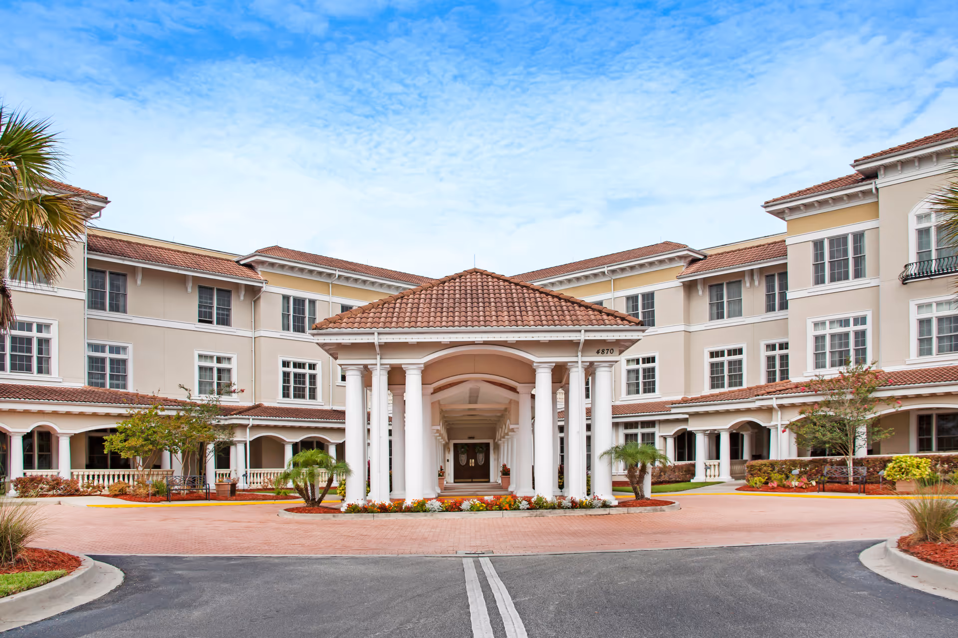Front entrance and porte-cochère of a three-story beige senior living building with tiled roofs, columns, and a landscaped circular driveway.