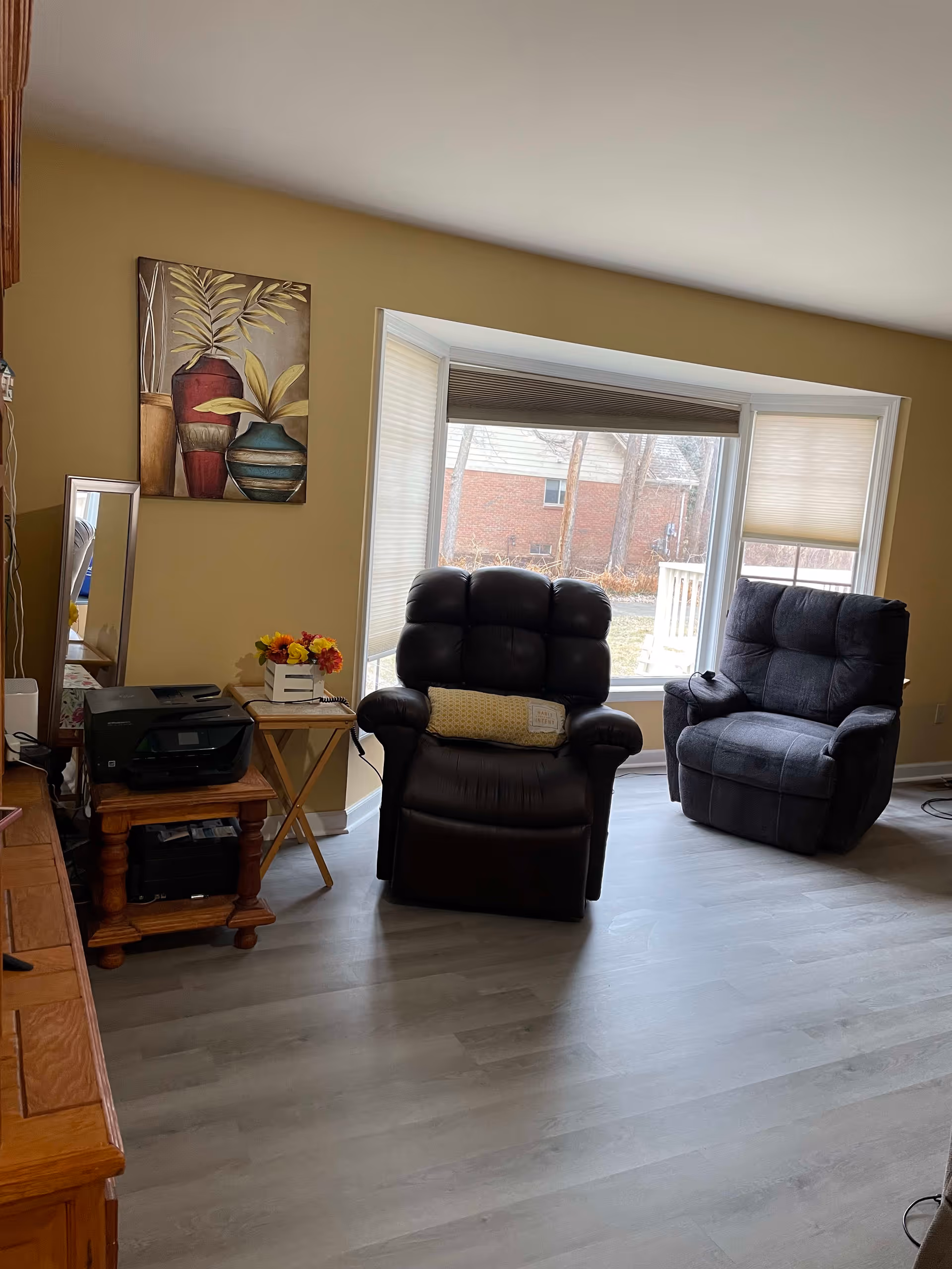 A cozy living room with two recliner chairs positioned near a large bay window with partially closed blinds. There is a small wooden table with a printer and a flower arrangement on it, and a decorative painting of vases on the wall. The floor is light-colored wood, and the walls are painted beige.