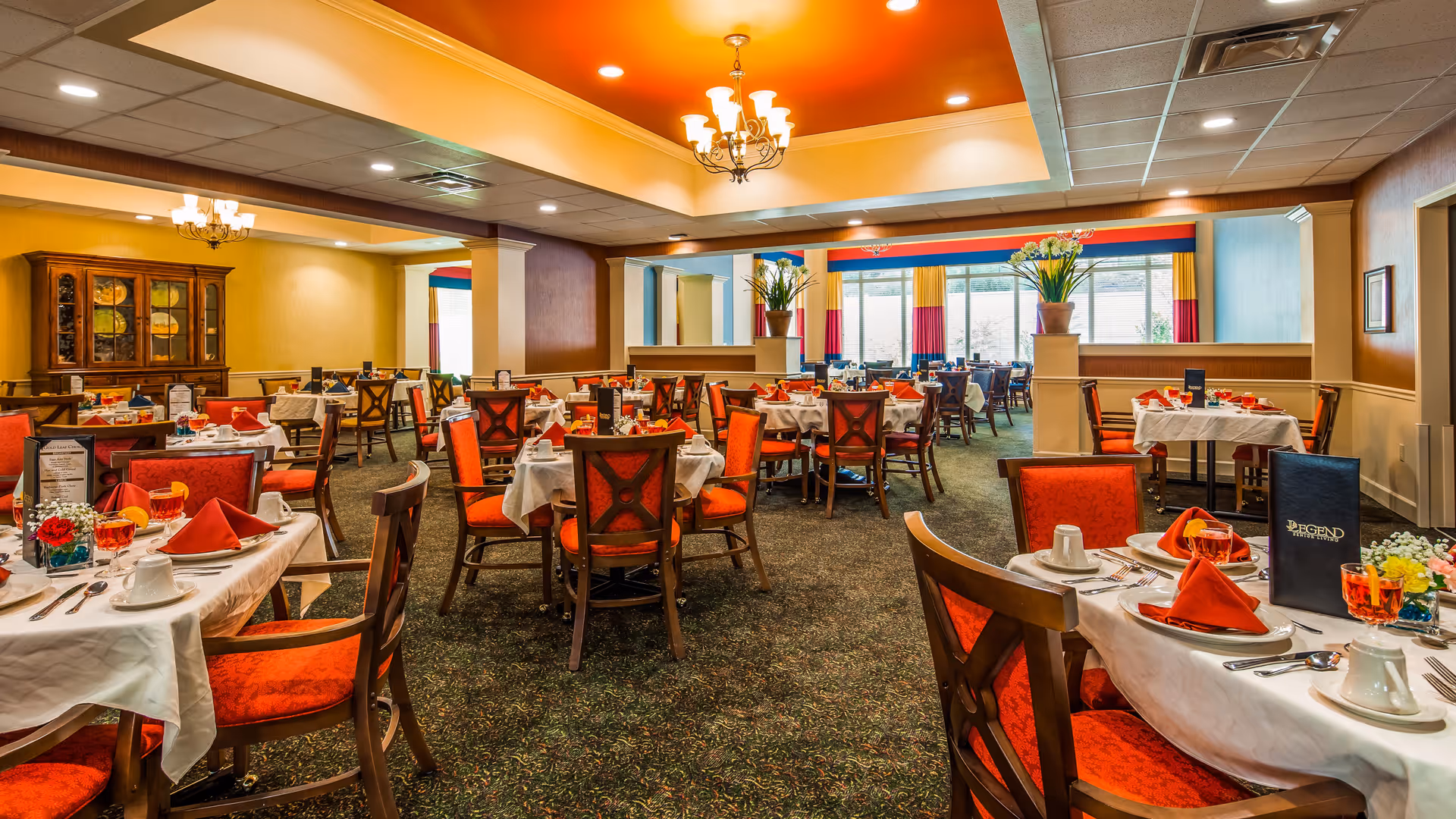 A spacious dining room with multiple tables set with white tablecloths, red napkins, plates, cups, and silverware. The chairs have red upholstery, and there are decorative plants on pillars. The room has warm lighting with chandeliers and large windows with colorful curtains allowing natural light in.