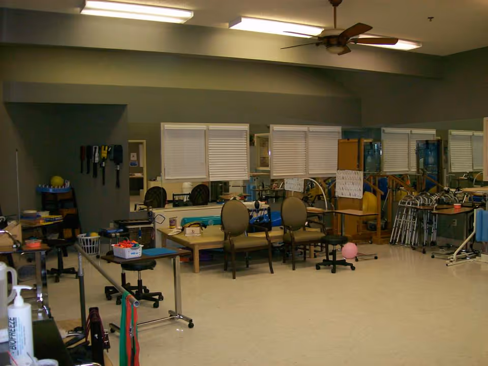 A physical therapy or rehabilitation room with exercise equipment, chairs, walkers, and therapy balls. The room has mirrors on the walls and various therapy tools and supplies are visible on tables and shelves.
