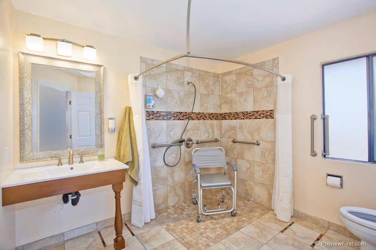 A spacious bathroom with beige tiled walls and floor, featuring a walk-in shower with a shower chair and grab bars. There is a large mirror above a wooden vanity with a sink, a green towel hanging on the wall, and a frosted window with grab bars next to a toilet.