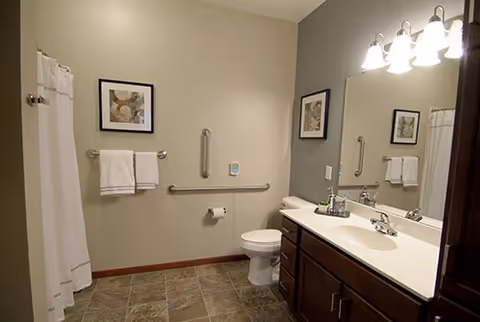 A clean and accessible bathroom with a white toilet, a white sink with a dark wood vanity, a large mirror, and a shower with a white curtain. There are two framed pictures on the walls, grab bars near the toilet, and two white towels hanging on a towel rack.