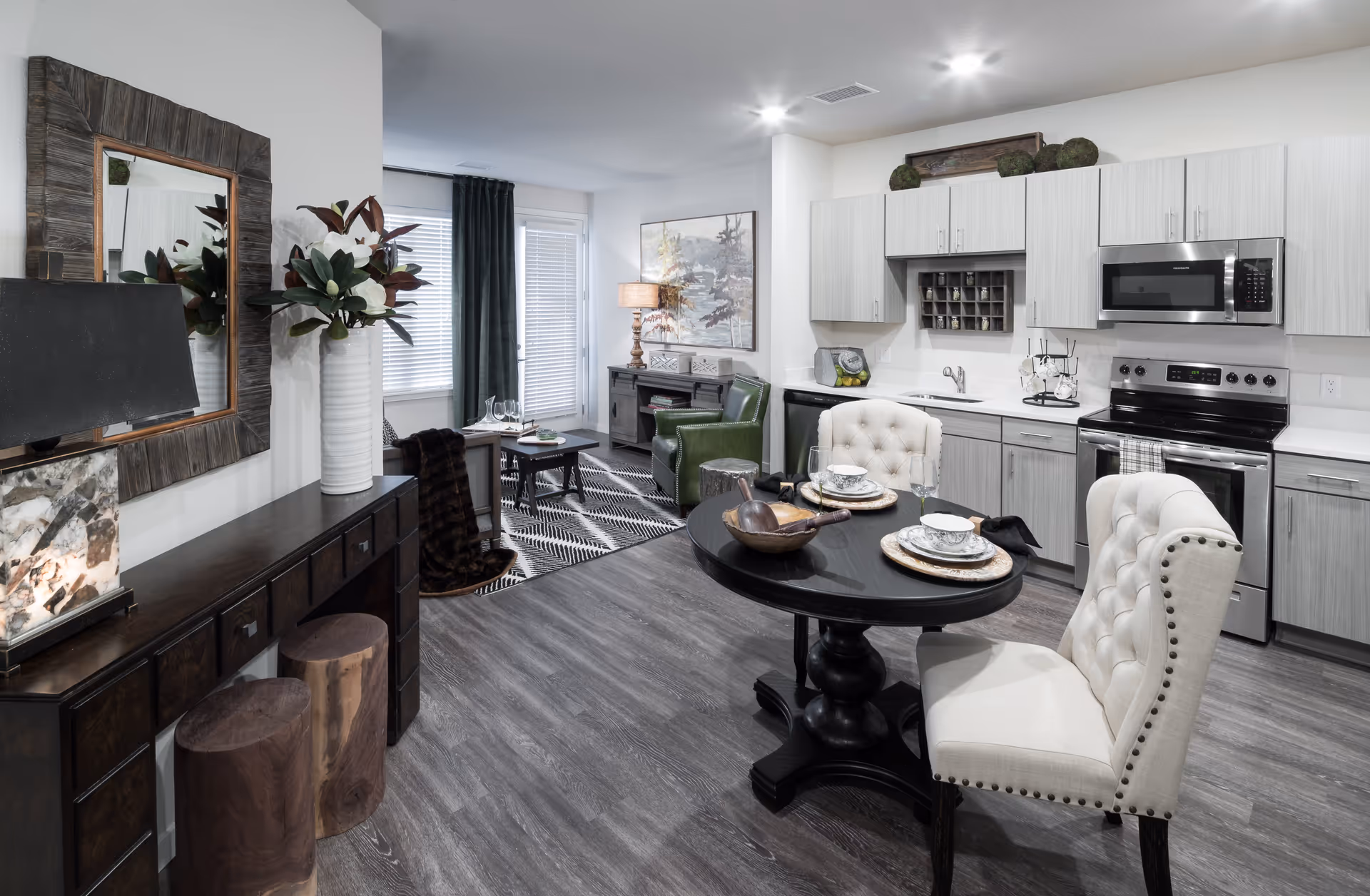 A modern open-concept living space featuring a kitchen with light wood cabinets, stainless steel appliances including a microwave and stove, and a small round dining table set for two with elegant dishware. Adjacent to the kitchen is a cozy living area with a green armchair, a patterned rug, a coffee table, and a sideboard with a lamp and decorative items. The room has wood flooring and large windows with dark curtains allowing natural light to enter.
