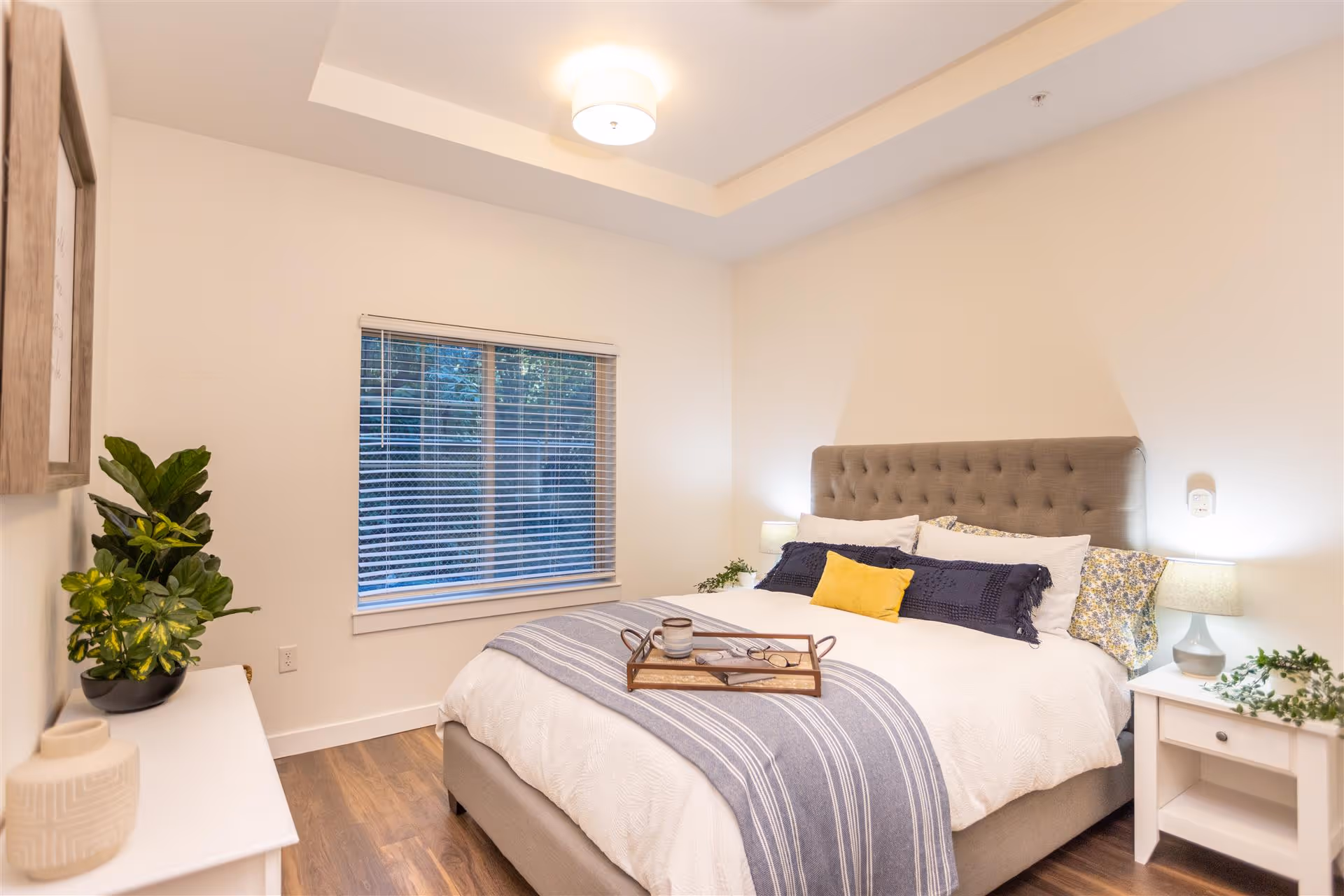 Bright, modern bedroom with a tufted upholstered bed dressed in white and blue linens, a decorative tray on the bed, nightstands, a potted plant and a window with blinds.