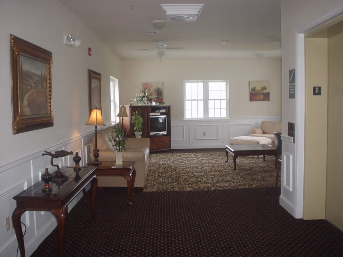 A well-lit interior common area with beige walls and carpeted floors. The room features a beige sofa, a wooden coffee table, a TV cabinet with a television, decorative plants, framed paintings on the walls, and a floor lamp. There is an elevator door marked with the number 2 on the right side of the image.