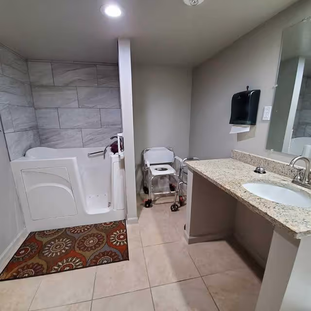 A bathroom with a walk-in bathtub on the left, a patterned rug in front of the tub, a white shower chair with wheels in the center, and a granite countertop with a sink and faucet on the right. A wall-mounted paper towel dispenser and a large mirror are above the sink.