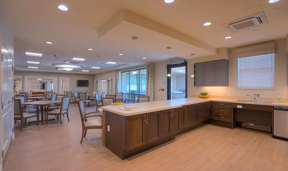 A spacious dining area with multiple tables and chairs arranged neatly. In the foreground, there is a large kitchen island with dark wooden cabinets and a light countertop. The kitchen area includes a sink, dishwasher, and upper cabinets. Large windows and glass doors allow natural light to fill the room. The ceiling has recessed lighting and air conditioning vents.