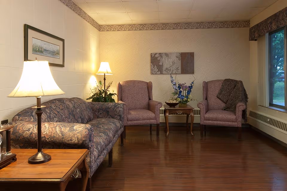 A cozy living room area with a floral patterned sofa on the left, two mauve upholstered armchairs with a small wooden table between them holding a floral arrangement. There are two lamps providing warm light, a framed picture on the wall above the sofa, and a large window on the right side showing greenery outside. The room has wooden flooring and light-colored walls.
