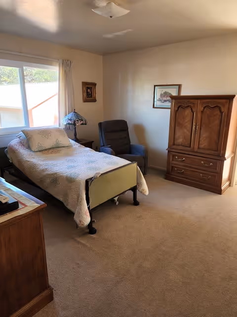 A simple bedroom with a single bed covered with a light-colored quilt and a pillow. Next to the bed is a small wooden nightstand with a decorative lamp. There is a cushioned armchair in the corner near the wall. A wooden armoire with drawers is positioned against the wall. The room has beige carpet and light-colored walls with two framed pictures hanging. A window with curtains allows natural light into the room.