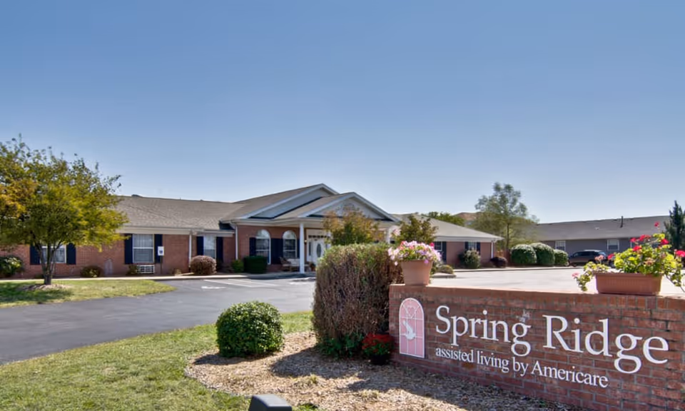 Exterior view of Spring Ridge Assisted Living facility showing a single-story brick building with a peaked roof, surrounded by trees and shrubs under a clear blue sky. In the foreground, there is a brick sign with the facility's name and flower pots on top.