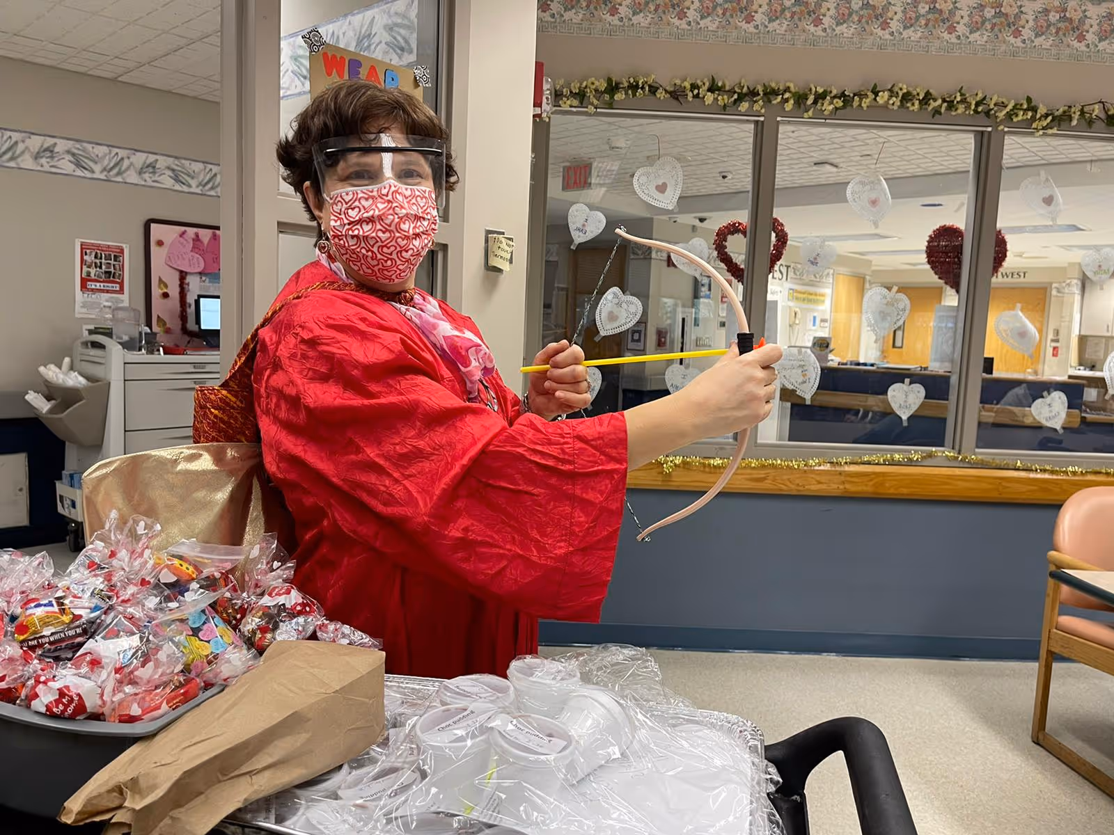 A person wearing a red outfit, a heart-patterned face mask, and a face shield is holding a toy bow and arrow indoors. The background shows a room decorated with heart-shaped decorations on the glass window, and there is a table with wrapped candy and plastic cups in the foreground.