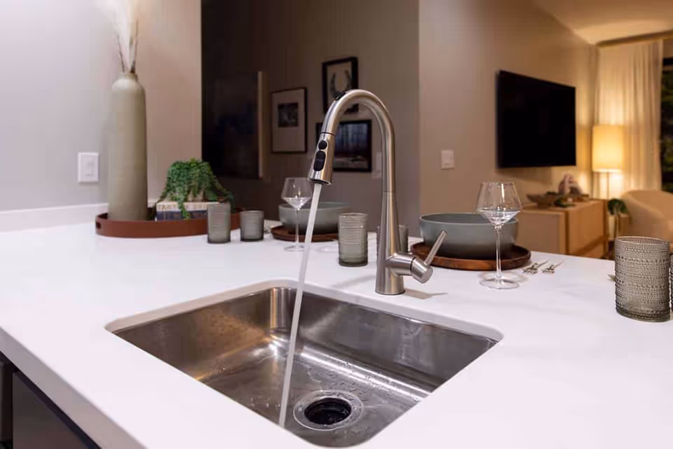 Modern kitchen sink with a stainless steel faucet running water, set in a white countertop. The countertop has decorative items including a tall vase with dried plants, a small green plant, glassware, and bowls. In the background, a living room area with a TV mounted on the wall and a lit floor lamp is visible.