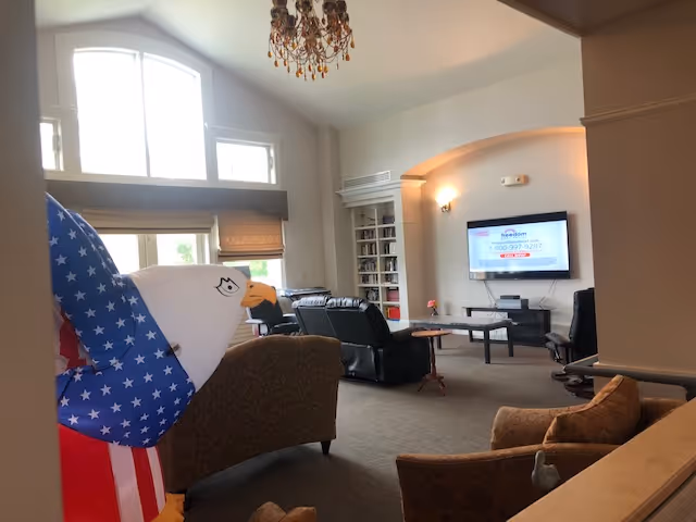 Interior view of a senior living facility common area with comfortable seating including armchairs and recliners, a large window letting in natural light, a mounted flat-screen TV on the wall, and a decorative eagle figure with an American flag design in the foreground.