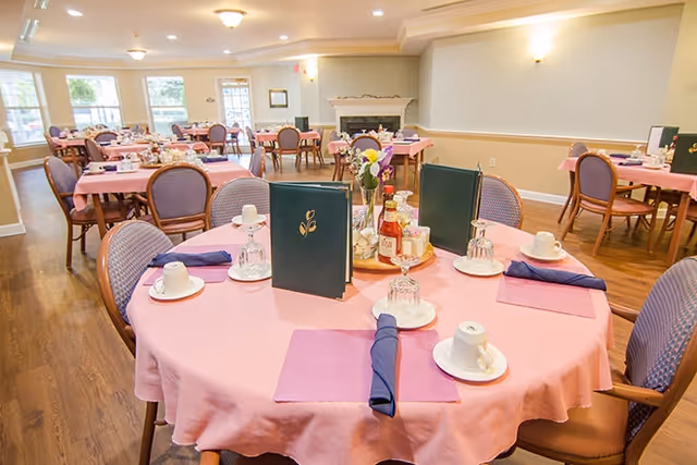 A dining room with multiple round tables covered in pink tablecloths, each set with cups, saucers, upside-down glasses, napkins, and menus. The room has wooden flooring, beige walls, and a fireplace in the background. Chairs with purple upholstery surround the tables.