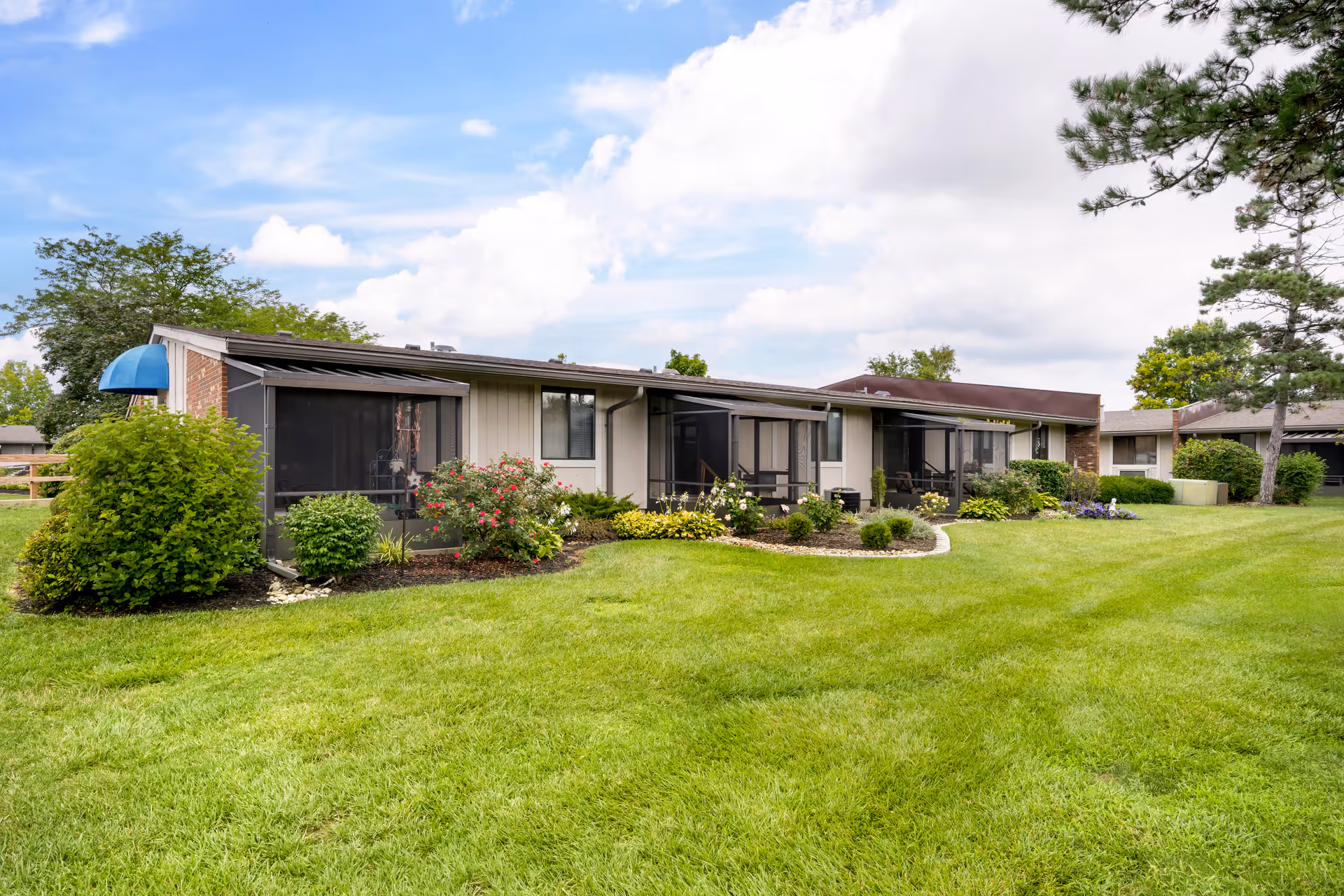 Exterior view of a single-story senior living facility building with screened-in patios, surrounded by well-maintained green grass, bushes, and flowering plants under a partly cloudy sky.