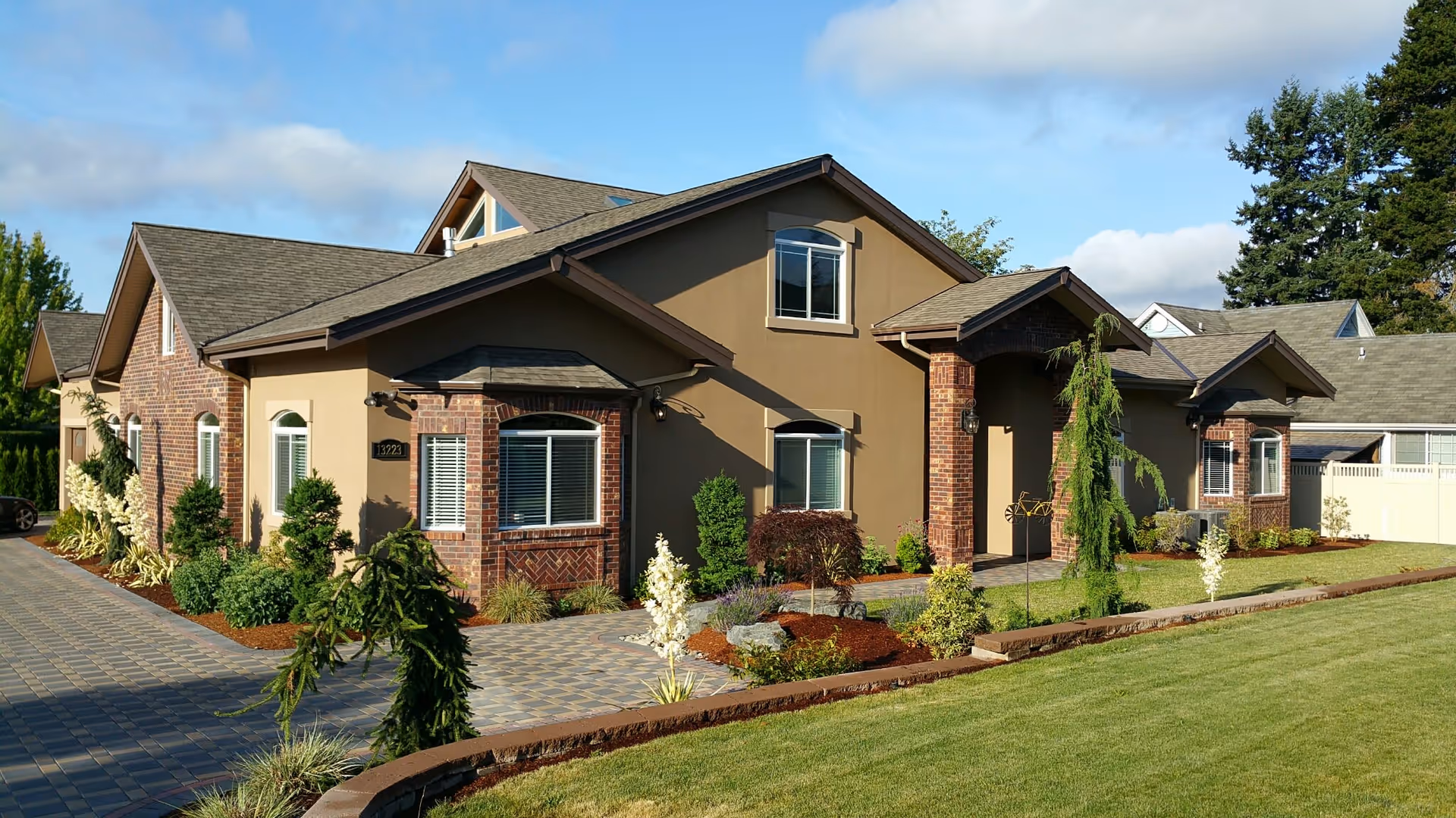 Exterior view of a single-story senior living facility building with a combination of brick and beige siding, multiple windows with white blinds, a paved walkway, and well-maintained landscaping including shrubs, small trees, and a green lawn under a partly cloudy blue sky.