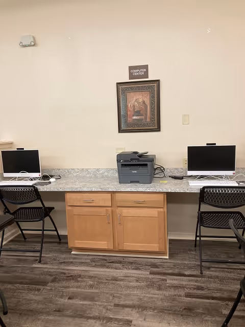 A computer center with two desktop computers on a long countertop with a printer in the middle. There are two black chairs in front of the computers. Above the printer, a framed picture is hung on the wall with a small sign above it that reads 'COMPUTER CENTER'. The floor has wood-patterned vinyl flooring.