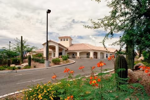 Exterior view of a single-story building with a tiled roof and arched entrance, surrounded by desert landscaping including cacti and flowering plants under a partly cloudy sky.
