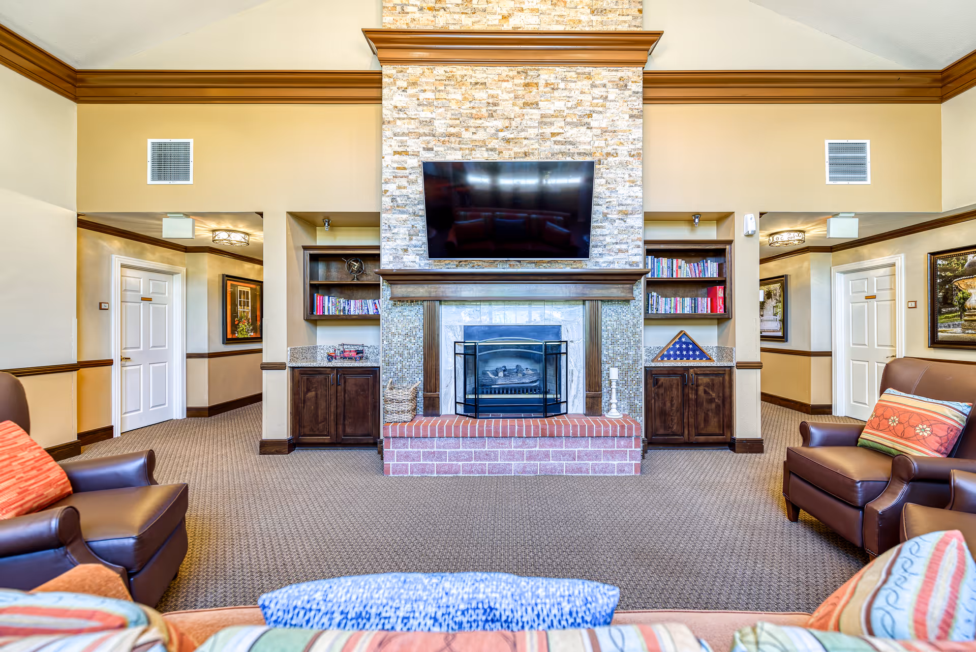 A cozy living room area in a senior living facility featuring a central fireplace with a stone facade and a mounted flat-screen TV above it. On either side of the fireplace are built-in wooden shelves filled with books and decorative items. Two brown leather armchairs with colorful cushions are positioned facing the fireplace, and a patterned sofa with multiple cushions is partially visible in the foreground. The room has beige walls with wooden trim and carpeted flooring.
