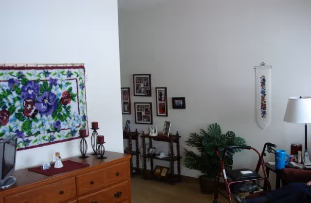 Interior view of a living area in Prairie Pines Assisted Living featuring a wooden dresser with decorative items, a floral wall hanging, a small table with framed photos, a potted plant, a walker, and a floor lamp.