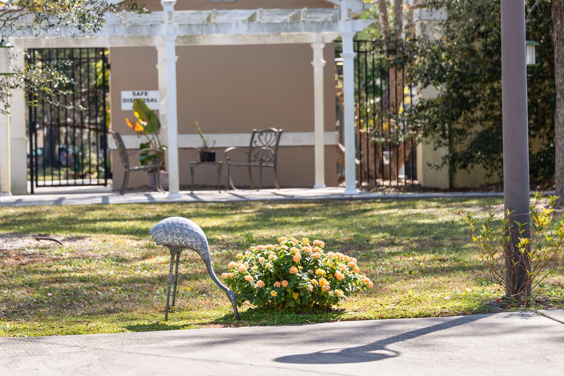 A metal heron lawn sculpture beside a flowering shrub in a grassy courtyard with a pergola and chairs in the background.