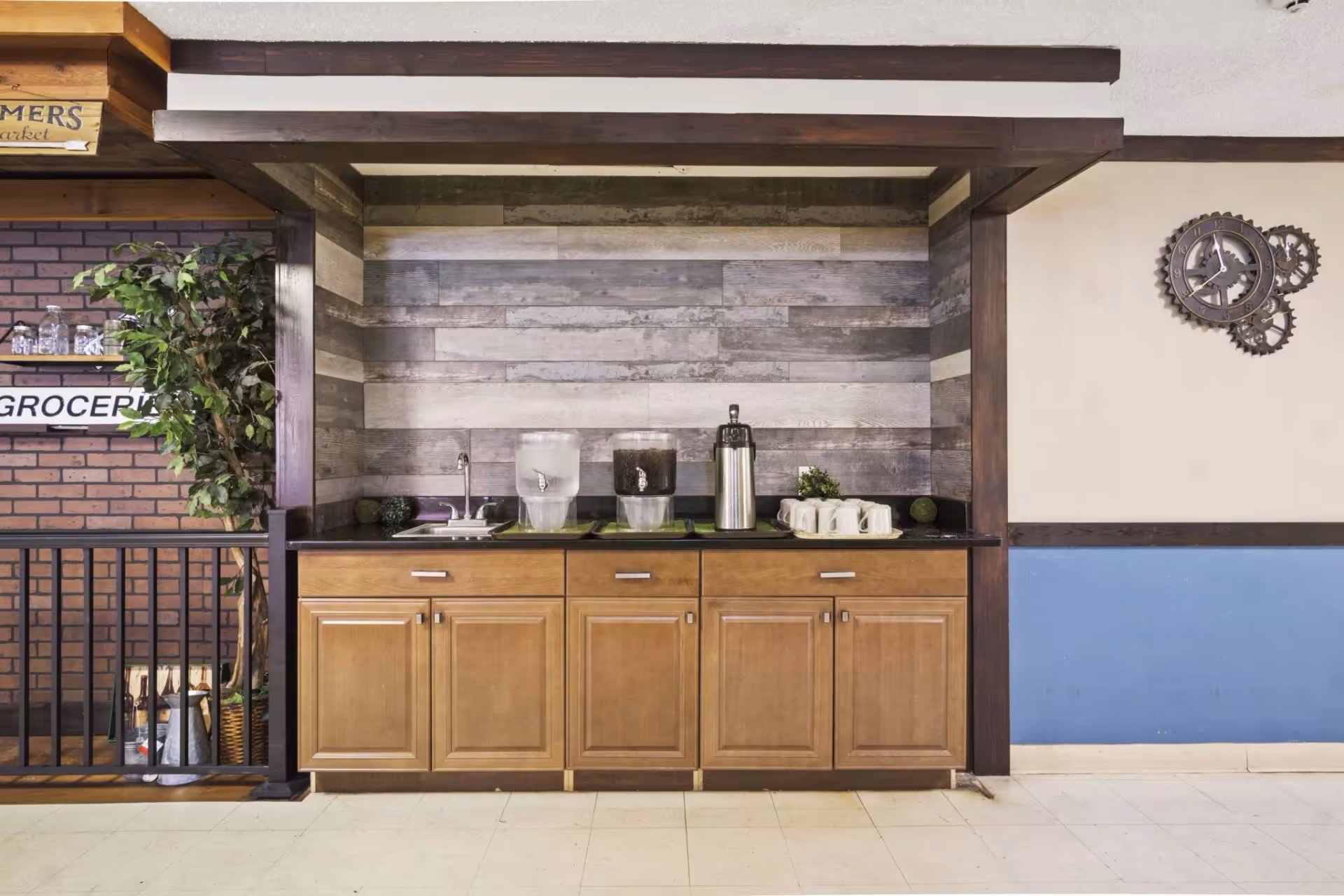 A beverage station with wooden cabinets, a small sink, two water dispensers, a coffee dispenser, and several white mugs on a tray. The background features a rustic wood panel wall and a decorative gear-shaped clock on the adjacent wall. There is a potted plant and a sign partially visible that reads 'GROCERY' on the left side.