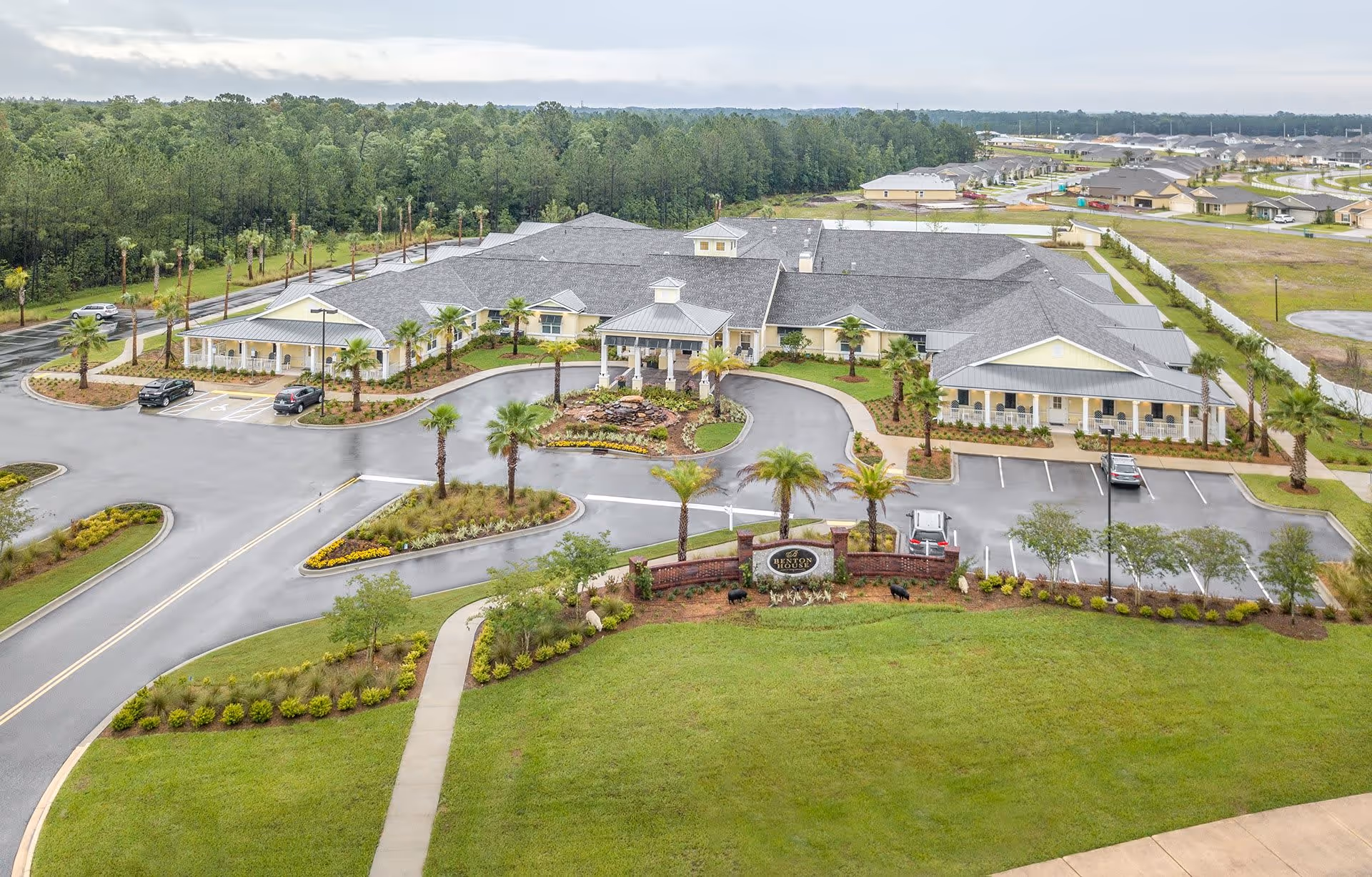 Aerial view of a large single-story senior living facility with a gray roof, surrounded by landscaped grounds with palm trees and parking spaces. The building has a covered entrance and is located near a wooded area with other buildings visible in the distance.