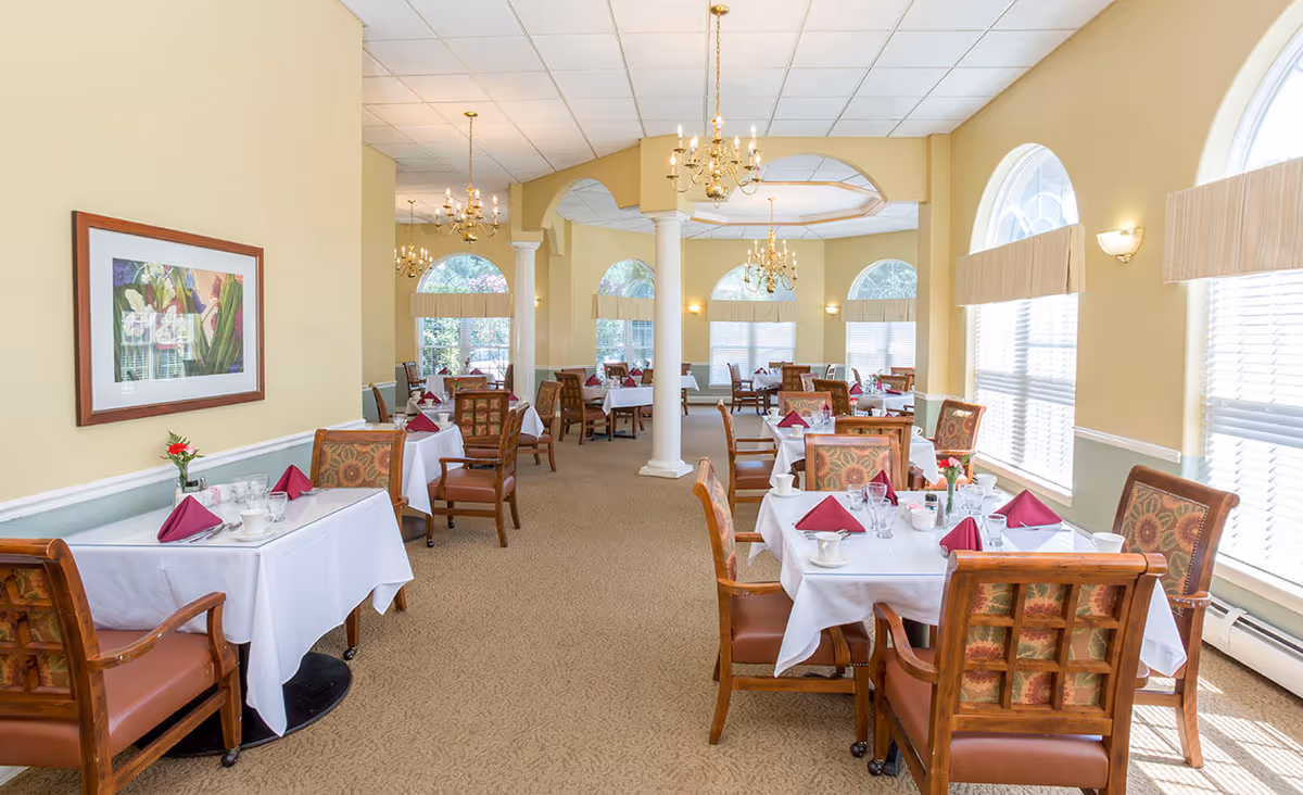 A bright and elegant dining room with multiple tables covered in white tablecloths, each set with burgundy folded napkins, glassware, and cups. The room features large arched windows with blinds, beige walls, carpeted floor, and decorative chandeliers hanging from the ceiling. Wooden chairs with patterned upholstery surround the tables, and a framed floral painting hangs on one wall.