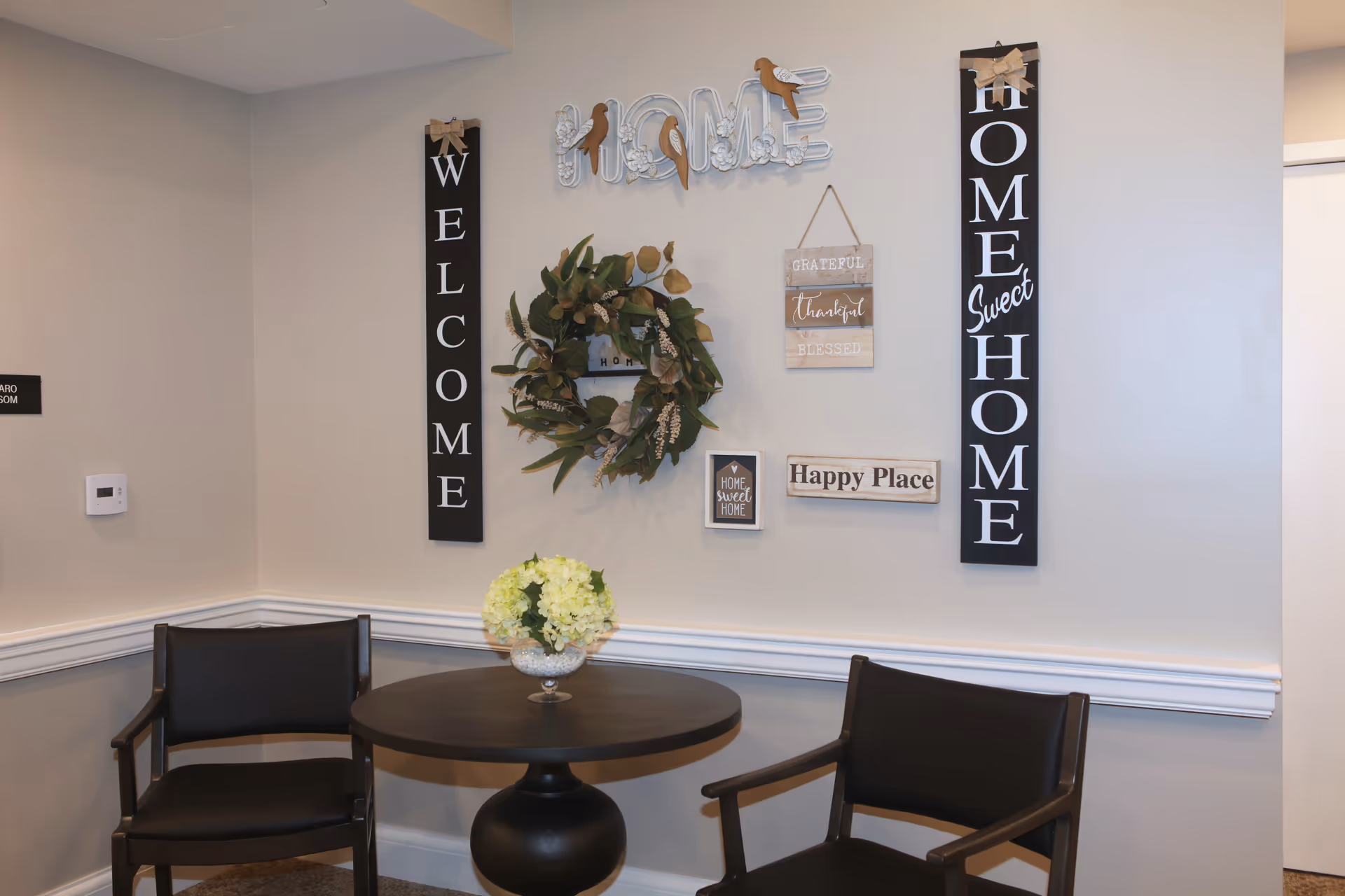 A cozy corner with two black chairs and a round black table with a glass vase holding white flowers. The wall behind features decorative signs including 'WELCOME', 'HOME', 'HOME Sweet HOME', 'GRATEFUL thankful BLESSED', and 'Happy Place', along with a green leafy wreath.