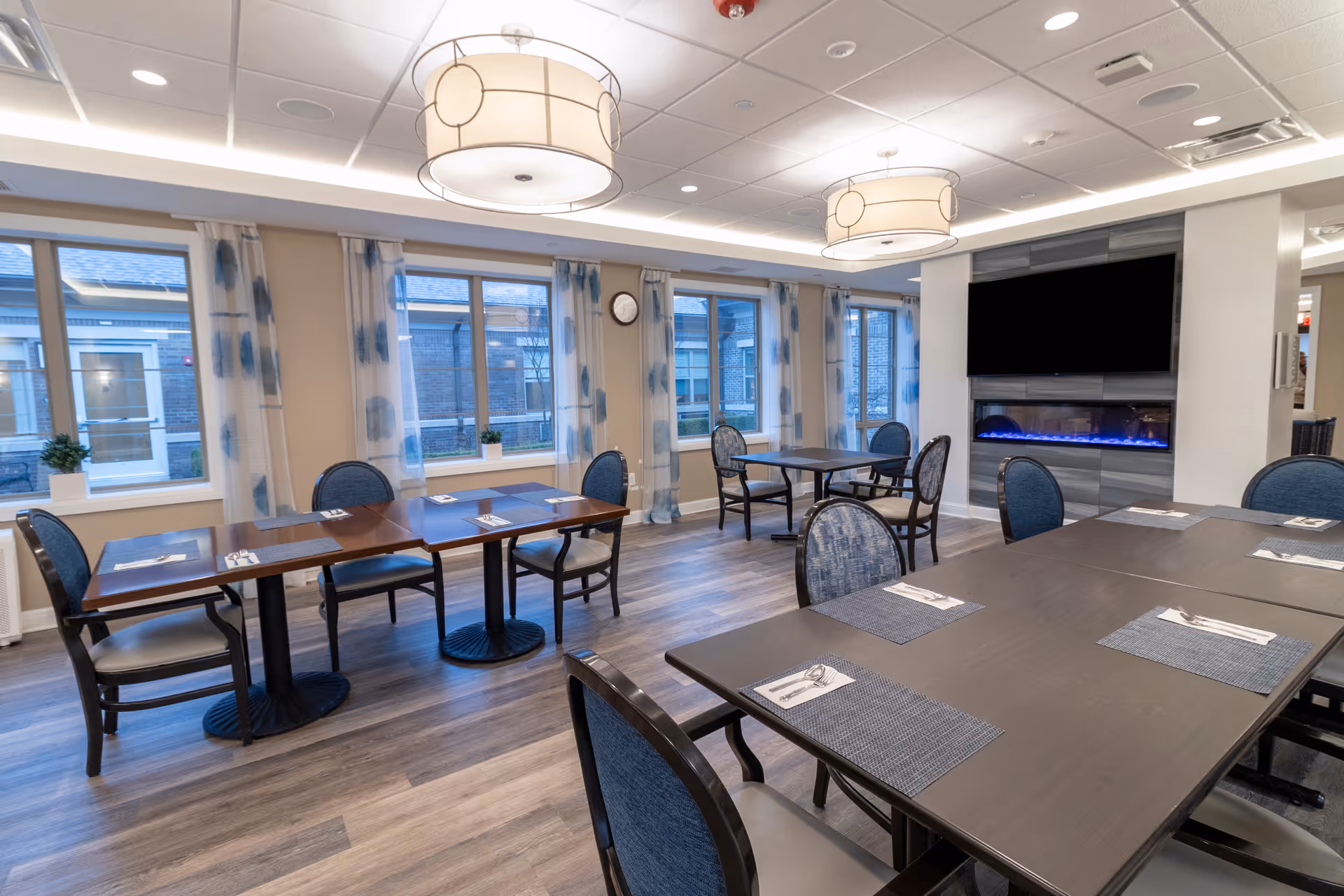 A bright dining room with several tables and chairs arranged neatly. The room features large windows with blue and white patterned curtains, modern ceiling lights, and a wall-mounted flat-screen TV above a sleek electric fireplace. The tables are set with placemats and silverware.