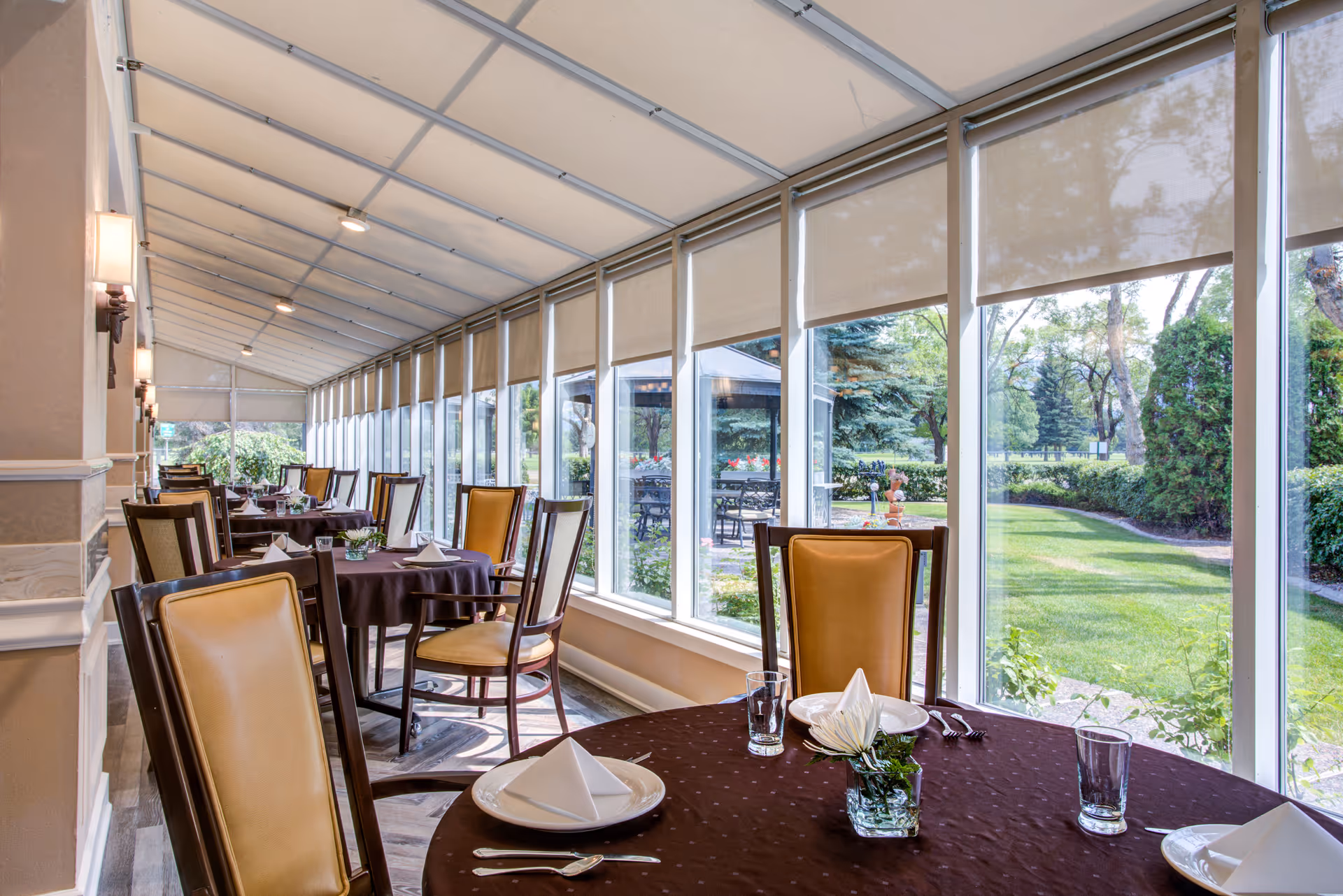 A bright dining area with multiple tables covered in dark brown tablecloths, each set with white plates, folded napkins, glasses, and silverware. The room has large windows with beige roller shades, offering a view of a green garden outside. The chairs have wooden frames with tan cushioned backs and seats.