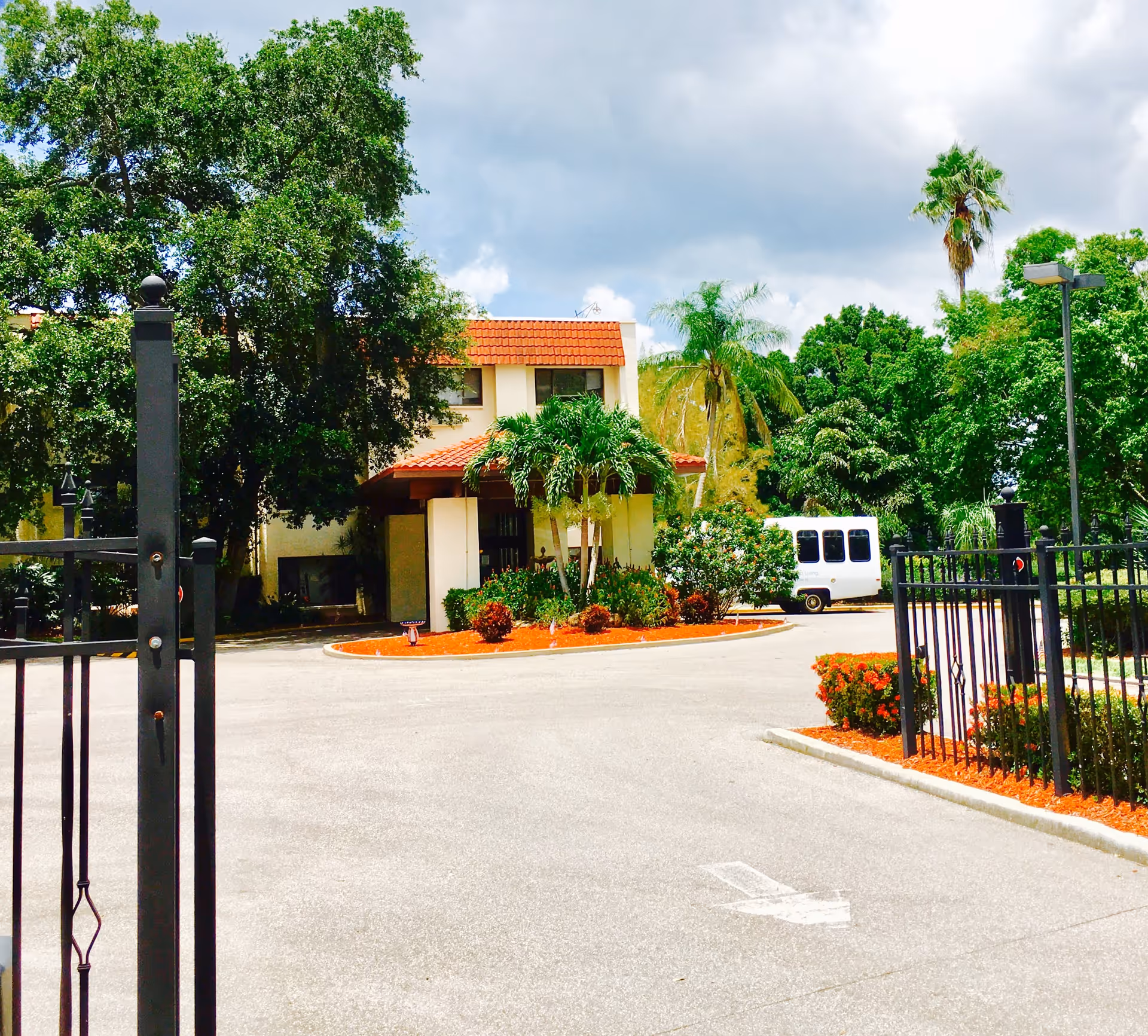 View of the entrance to a building with a red-tiled roof surrounded by lush green trees and landscaping, including palm trees and bushes. A white van is parked near the building, and a black metal gate is partially visible in the foreground.