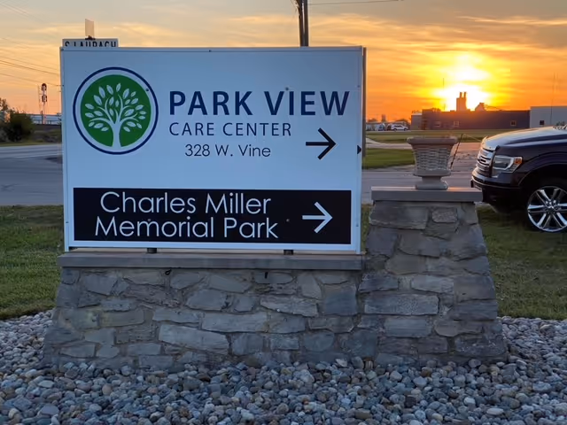 A stone signpost with a white sign displaying the text 'PARK VIEW CARE CENTER 328 W. Vine' and an arrow pointing right, along with 'Charles Miller Memorial Park' and an arrow pointing right. The sign is set on a bed of small rocks with a sunset and a parked vehicle in the background.